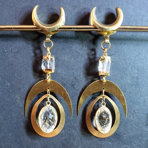 Puede incluir: Un par de pendientes de tono dorado con un diseño de media luna y una piedra de cristal transparente. Los pendientes cuelgan de un aro dorado.