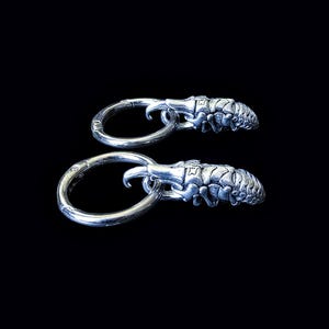 Könnte beinhalten: Zwei silberfarbene Creolen mit detailliertem, insektenartigem Design. Jeder Ohrring hat einen Klappbügel und ein dekoratives Element, das einem segmentierten Körper mit einem gebogenen, klauenartigen Anhang ähnelt. Der Hintergrund ist schwarz.