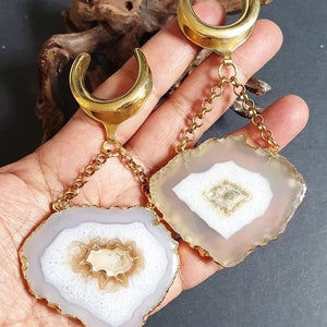 Puede incluir: Un par de expansores de oreja dorados en forma de media luna con colgantes de piedra de ágata de forma irregular. Las piedras de ágata tienen un centro blanco, rodeado de bandas marrones y grises, y están bordeadas con oro. Los expansores están conectados a los colgantes por cadenas doradas.