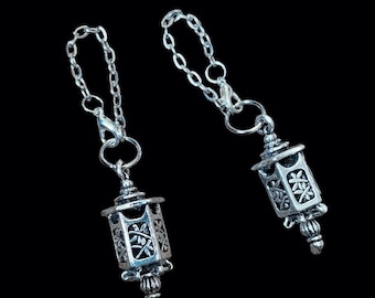 May include: Two silver-toned metal charms shaped like miniature lanterns, each featuring intricate cut-out floral patterns on the sides and a small dangling finial at the bottom. Each lantern charm is attached to a silver-colored metal chain link.