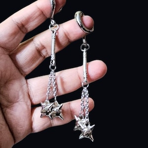 以下が含まれることがあります： ユニークなデザインのシルバーカラーのイヤリング。各イヤリングは、湾曲した上部、長く細い中央部分、とげのある星型のチャームを吊り下げているチェーンが特徴です。イヤリングは暗い背景に表示されています。
