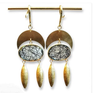 May include: Gold-toned dangle earrings with a crescent moon shape, oval-shaped stone with black inclusions, and textured leaf-shaped pendants. The earrings are suspended from a gold bar.