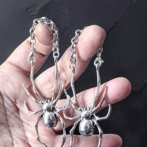 May include: A pair of silver-toned spider-shaped earrings, each featuring a metallic spider body with eight articulated legs attached to a circular link chain with a lobster claw clasp, creating a dangling design with a polished finish.