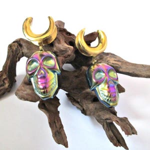 May include: A pair of gold-colored ear gauges with iridescent skull charms. The gauges are made of stainless steel and the skulls are made of glass.