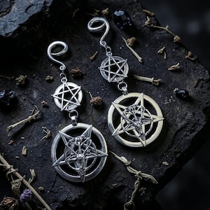 May include: Silver earrings with a pentagram design. Each earring has a curved top, a smaller pentagram, and a larger circular pendant with a detailed pentagram and a goat-like figure. The earrings are presented on a dark, textured surface.