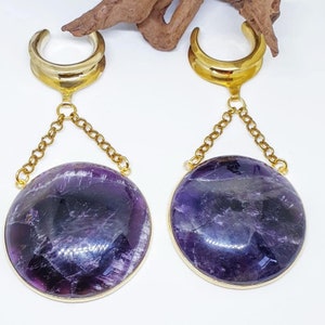 May include: A pair of gold-tone ear gauges with amethyst stone pendants. The gauges are shaped like a crescent moon and are connected to the stones by a gold chain.