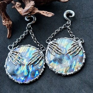 Puede incluir: Un par de pendientes de plata con forma de mano de esqueleto con detalles de piedra de labradorita iridiscente. Los pendientes están unidos a una cadena de plata con cierre de gancho.