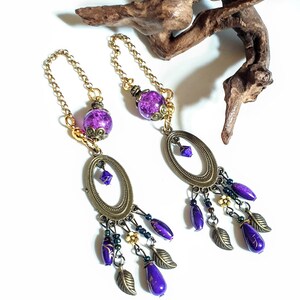 May include: Purple and gold dangle earrings with a large oval charm and multiple purple beads. The earrings are hanging from a gold chain.