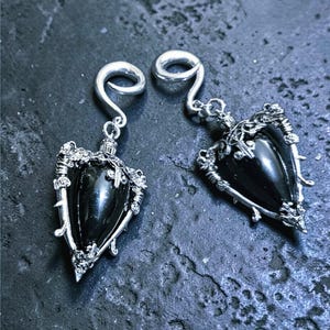 Peut inclure: Une paire de boucles d'oreilles argentées avec une pierre centrale sombre en forme de goutte. Les boucles d'oreilles présentent des détails complexes et ornés autour de la pierre et un crochet incurvé pour les oreilles percées. Le design a une esthétique gothique ou fantastique.