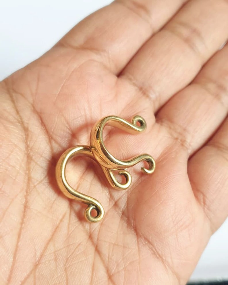 May include: Gold-toned metal nose ring with a unique, intricate design. The ring is shaped like a double loop with a smaller loop at the end of each larger loop.
