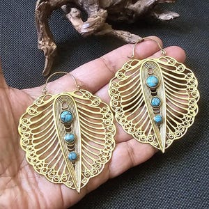 Può includere: Un paio di orecchini in filigrana color oro a forma di foglia, ciascuno con tre pietre color turchese. Gli orecchini hanno un motivo traforato dettagliato e una chiusura a gancio. Gli orecchini sono esposti su una mano.
