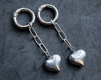May include: A pair of silver earrings. Each earring has a polished heart pendant hanging from a chain. The chain is attached to a circular clasp. The background is dark grey.