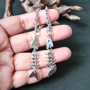 Könnte beinhalten: Ein Paar silberne Fischgräten-Ohrringe mit einem Kettenglied-Design. Die Ohrringe hängen an einer silbernen Kette.