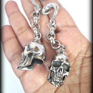 Puede incluir: Un par de pendientes de calavera plateados. Cada pendiente presenta un colgante de calavera detallado suspendido de una cadena y un gancho curvo. Las calaveras tienen cuencas de los ojos, narices y dientes visibles, y están hechas de metal brillante.