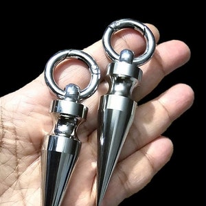 Puede incluir: Dos colgantes de llavero de metal plateado con punta y un anillo en la parte superior. Los colgantes tienen forma de cono con una punta redondeada.