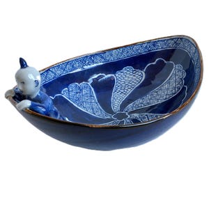 May include: A blue and white ceramic bowl with a decorative floral pattern. A small figurine of a baby is attached to the side of the bowl. The bowl has a dark brown rim and is set against a white background.