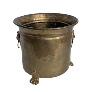 Puede incluir: Una jardinera de latón martillado con un borde ancho y tres patas de garra. La jardinera tiene dos asas con cabeza de león y una superficie texturizada. Esta pieza decorativa es ideal para uso en interiores o exteriores.