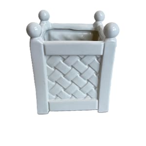 Könnte beinhalten: Ein quadratischer, weißer Keramik-Pflanztopf mit einem geflochtenen Korbmuster an den Seiten. Der Pflanztopf hat eine rechteckige Öffnung und ist mit vier kleinen, runden Knäufen versehen. Das Design ist schlicht und klassisch.