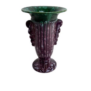 Peut inclure: Un vase décoratif en céramique avec un bord vert foncé et un corps violet foncé. Le vase présente un design cannelé et des détails de visage sculptés de chaque côté. La base est ronde et le vase mesure environ 25 cm de haut.