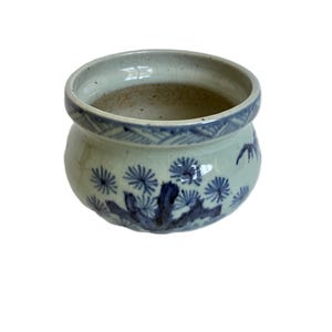 Pode incluir: Pequeno vaso redondo de cerâmica branca com padrões florais e geométricos azuis. O vaso tem uma abertura larga e uma superfície ligeiramente texturizada. Os desenhos azuis são pintados à mão, dando-lhe um aspeto artesanal único.