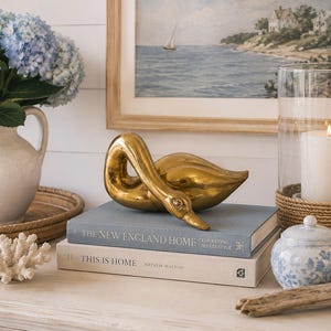 Pode incluir: Uma escultura de cisne dourado repousa sobre dois livros, "The New England Home" e "This is Home". Um vaso branco com hortênsias azuis está à esquerda. Uma vela acesa numa lanterna de vidro e um pote azul e branco também estão presentes.