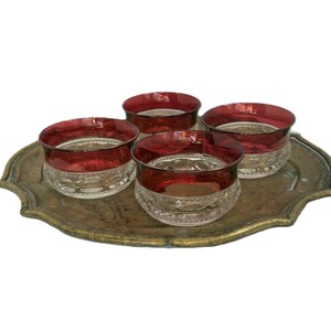 Könnte beinhalten: Ein Set aus vier kleinen Glasschalen mit tiefroten Rändern und klaren, strukturierten Böden mit einem sich wiederholenden kreisförmigen Muster, angeordnet auf einem ovalen, verwitterten messingfarbenen Metalltablett mit gewelltem Rand.