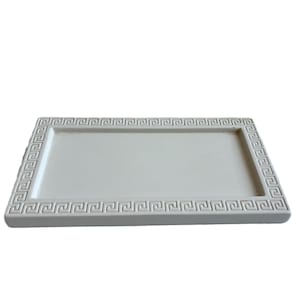 May include: A rectangular, off-white tray with a Greek key design border. The tray is made of a solid material and has a slightly raised edge. The design is a repeating pattern around the perimeter.