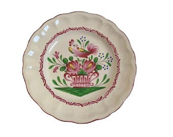 May include: A cream-colored ceramic plate with a scalloped edge and a hand-painted design. The center features a pink and green rooster perched above a floral arrangement in a pink urn, surrounded by a decorative red border along the rim.