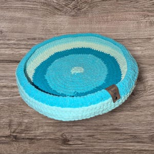May include: A circular pet bed, hand-crocheted in varying shades of blue and turquoise. The bed has a raised edge and a small, brown leather tag. The bed is designed for small pets, and is made of soft yarn.
