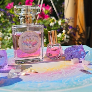 May include: A clear glass perfume bottle and a smaller bottle with gold caps, labeled "Andara Crystal Magical Aura Spray." The bottles are filled with pink liquid and surrounded by amethyst and quartz crystals. The background features colorful flowers.