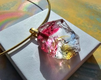 May include: A gold chain necklace with a large, clear, faceted pendant. The pendant is filled with pink, yellow, and white resin.