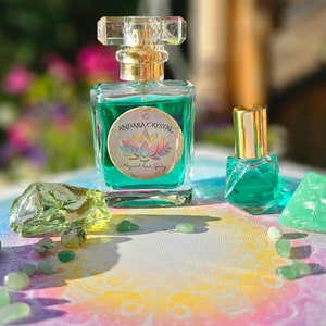 Puede incluir: Dos botellas de l&iacute;quido verde con tapas y etiquetas doradas. La botella m&aacute;s grande tiene una etiqueta que dice "Andara Crystal Magical Aura Spray". Las botellas est&aacute;n rodeadas de cristales verdes sobre una superficie blanca con un patr&oacute;n de arco&iacute;ris.