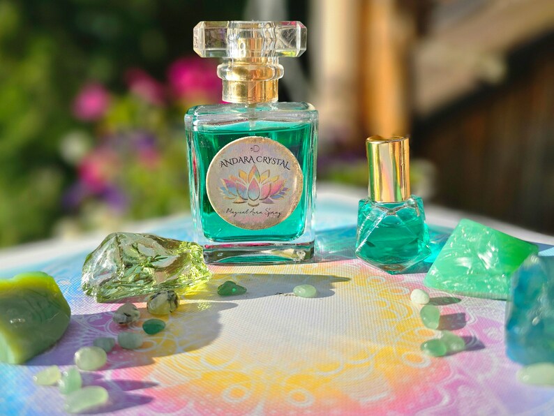 Puede incluir: Dos botellas de l&iacute;quido verde con tapas y etiquetas doradas. La botella m&aacute;s grande tiene una etiqueta que dice "Andara Crystal Magical Aura Spray". Las botellas est&aacute;n rodeadas de cristales verdes sobre una superficie blanca con un patr&oacute;n de arco&iacute;ris.