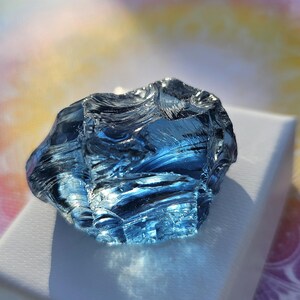 Puede incluir: Un gran cristal azul facetado con una superficie rugosa y texturizada. El cristal está sobre una superficie blanca.