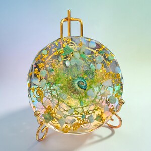 May include: A round, clear resin art piece with embedded green, blue, and gold elements, including a spiral shell. The artwork is displayed on a small, gold-colored easel with a three-legged base. The piece has a shimmering, iridescent quality.