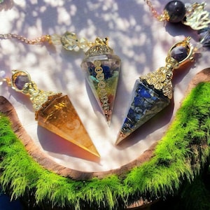 May include: Three gold-toned crystal pendulums with different colored crystals. The pendulums are hanging from chains and are on a wooden surface with green moss.