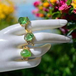 Könnte beinhalten: Drei goldene Ringe mit grünen Edelsteinen und kleinen Diamanten. Die Ringe befinden sich an einer weißen Mannequin-Hand.