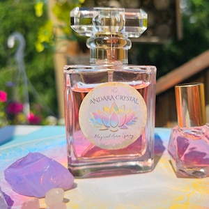 May include: A clear glass bottle with a gold cap and a pink label that reads "3D Andara Crystal Magical Aura Spray" with a pink lotus flower design. The bottle is sitting on a table with pink and white crystals.