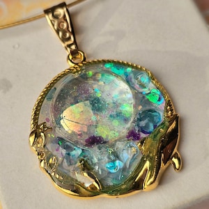 Può includere: Una collana con un ciondolo rotondo in resina trasparente riempito di glitter iridescente e accenti blu e verdi. Il ciondolo è circondato da una cornice dorata con un design decorativo.