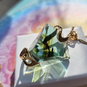 May include: A gold necklace with a clear, geometric pendant. The pendant has a gold-coloured metal frame and contains green and brown inclusions. The necklace is displayed on a white surface, with a colourful background.