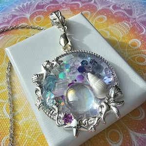 Può includere: Una collana con pendente argentato con un centro in resina trasparente riempito di glitter blu, viola e iridescenti, piccoli cristalli e una conchiglia bianca. Il pendente è circondato da una cornice argentata con stelle marine e un bordo a forma di corda. La collana è su una scatola bianca.
