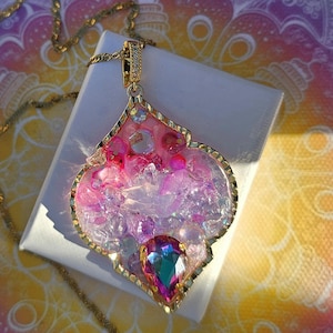 May include: A gold-coloured necklace with a pendant featuring a pink and purple resin design, accented with clear crystals and a large, teardrop-shaped gemstone. The pendant is set on a gold chain.