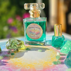 Puede incluir: Dos botellas de l&iacute;quido verde con tapas doradas y etiquetas que dicen "Andara Crystal Magical Aura Spray". Las botellas est&aacute;n sobre una superficie blanca con piedras verdes esparcidas a su alrededor.