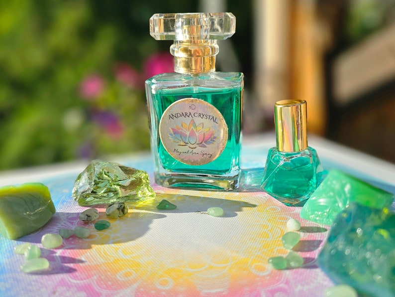 Puede incluir: Dos botellas de l&iacute;quido verde con tapas doradas y etiquetas que dicen "Andara Crystal Magical Aura Spray". Las botellas est&aacute;n sobre una superficie blanca con piedras verdes esparcidas a su alrededor.