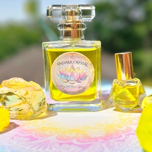 May include: Two glass bottles of yellow liquid with gold caps. The larger bottle has a label that reads "Andara Crystal Magical Aura Spray". The bottles are sitting on a colorful surface with yellow crystals.