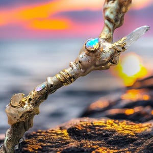May include: A close-up of a fantasy staff with a gold and silver finish. The staff has a clear crystal tip and is decorated with iridescent gemstones. The background shows a sunset over the sea, with the staff resting on a dark, textured surface.
