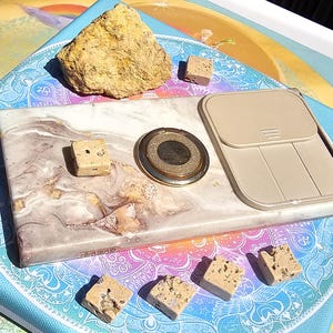 May include: A display of natural stone and decorative objects. Features a large, rough yellow rock, small stone cubes, a marble-effect tray, a gold-edged circular item, and a beige phone case-style object. The items are arranged on a colourful, patterned surface.