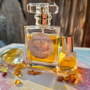 May include: Two glass bottles of golden yellow liquid with gold caps. The larger bottle has a label that reads "Andara Crystal Magical Aura Spray". The bottles are surrounded by small, clear and amber-colored crystals on a colorful surface.