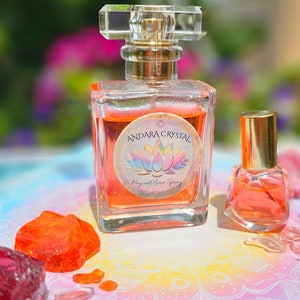 May include: Two glass bottles of orange-colored liquid with gold caps. The larger bottle has a label that reads "3D Andara Crystal Magical Aura Spray". The bottles are sitting on a colorful surface with several orange and pink crystals.
