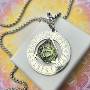 May include: A silver pendant necklace featuring a circular pendant with engraved runes and a central, faceted green crystal. The pendant hangs from a silver chain, displayed on a white surface.